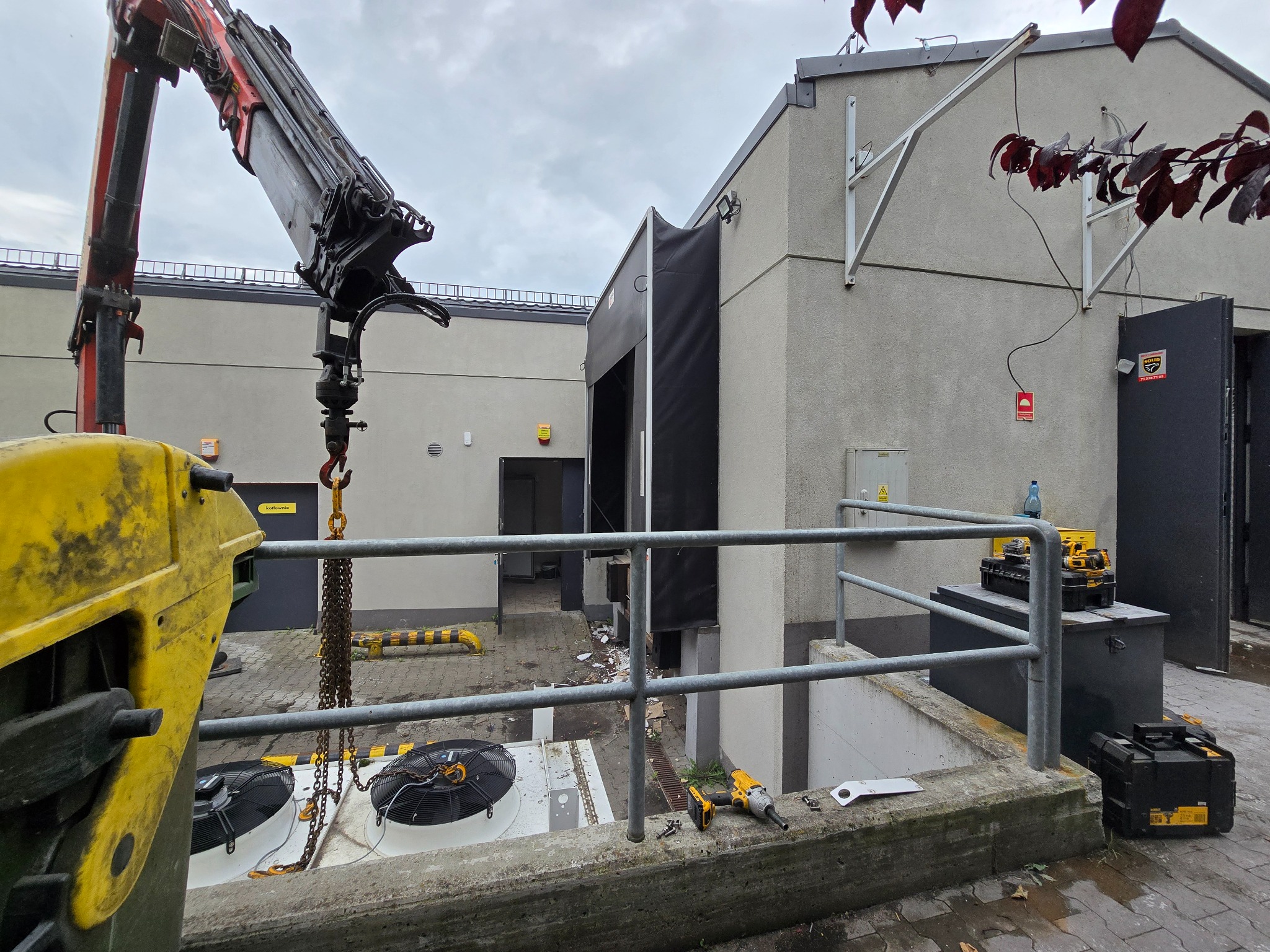 Żuraw przenosi agregaty chłodnicze na zewnątrz budynku. Widoczne narzędzia i skrzynki narzędziowe. Szaro-ponura pogoda.