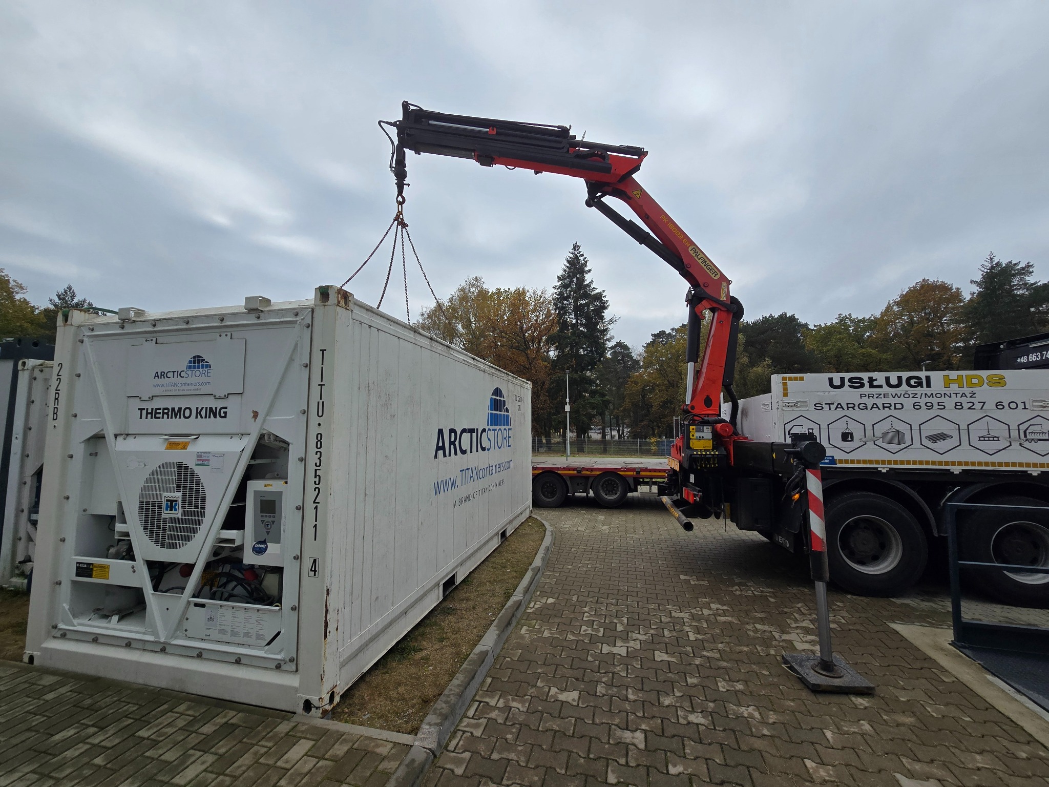 Ciężarówka HDS z dźwigiem podnosi biały kontener chłodniczy ArcticStore na tle pochmurnego nieba. Widoczny numer kontenera i logo Thermo King.