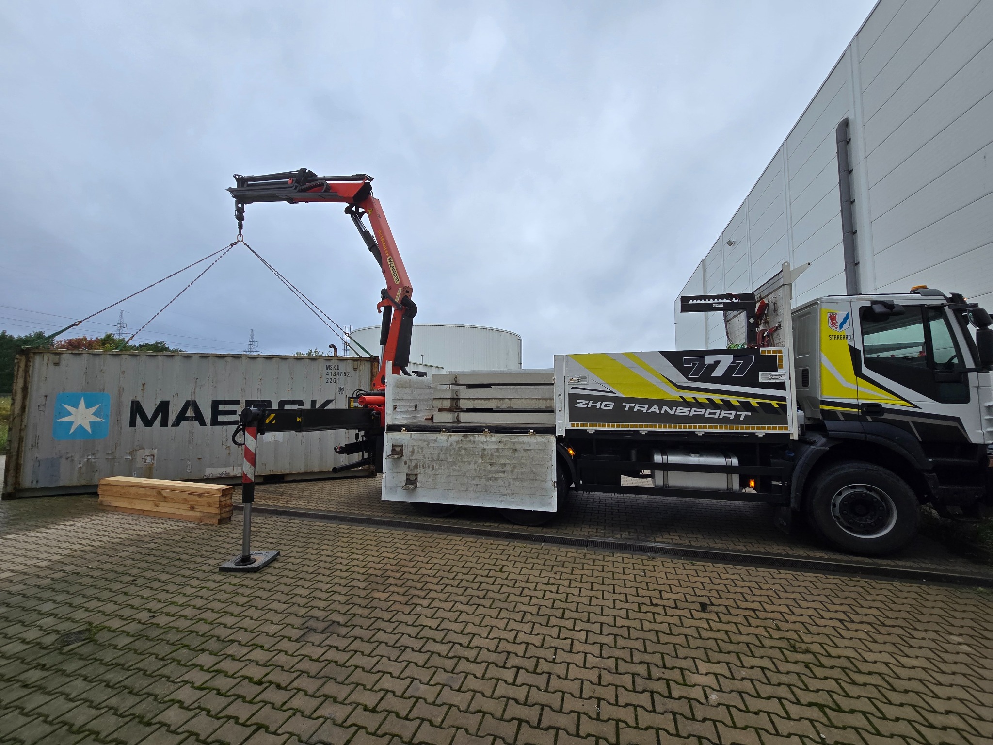Ciężarówka z dźwigiem Palfinger podnosi kontener Maersk na tle magazynu i zbiorników. Widoczne deski i kostka brukowa.