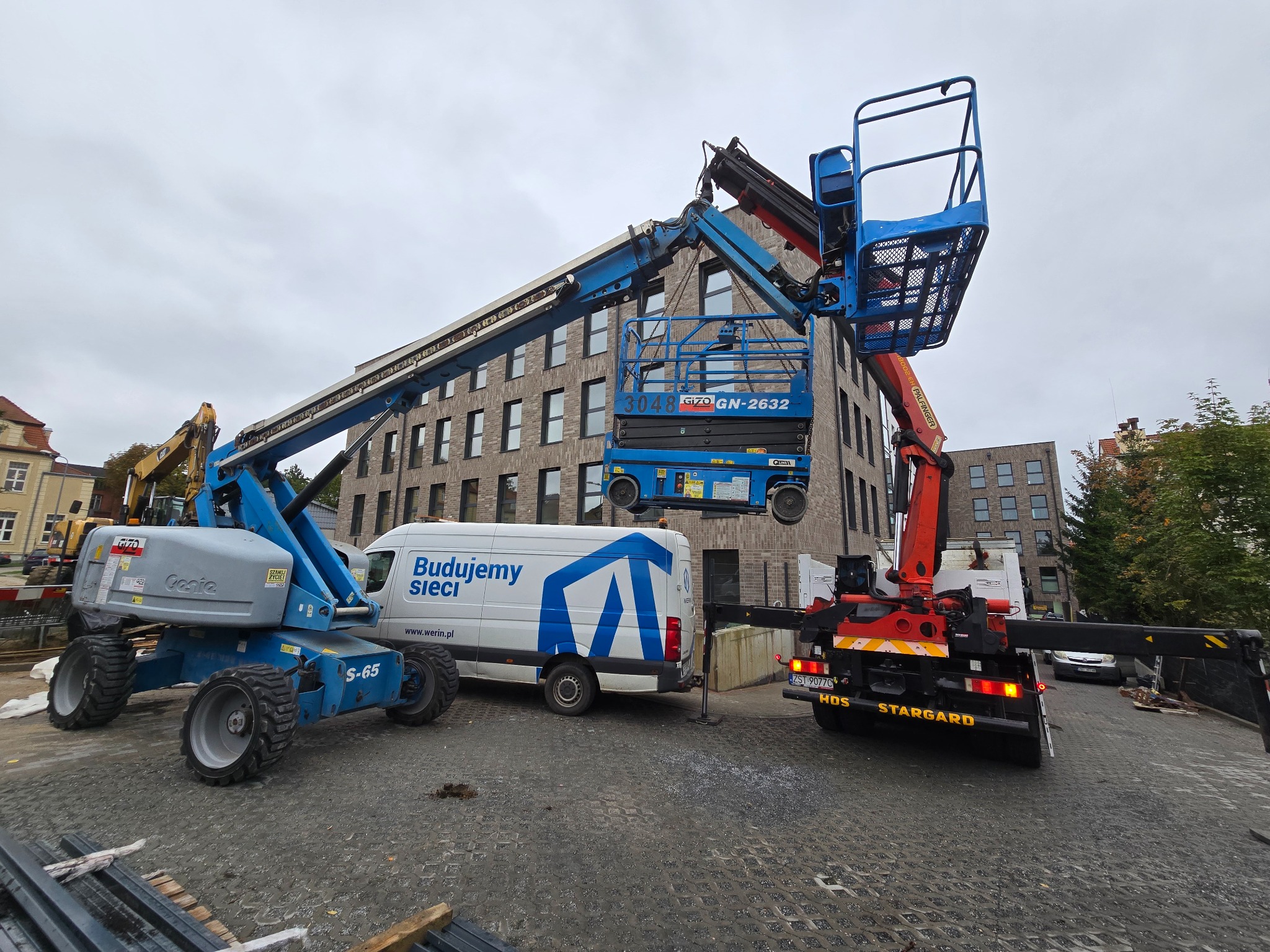 Dwie podnośniki koszowe (Genie S-65 i HDS Stargard) na placu budowy, z budynkiem w tle i furgonetką 'Budujemy sieci'.