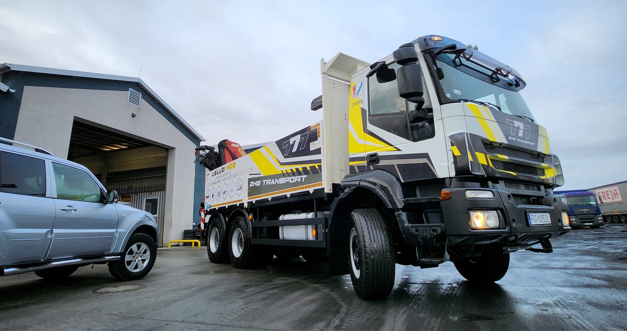 Żółto-czarno-biała ciężarówka z HDS ZHG Transport na mokrym placu, obok srebrny SUV, w tle budynek z otwartą bramą i inne ciężarówki. Widok z dołu.