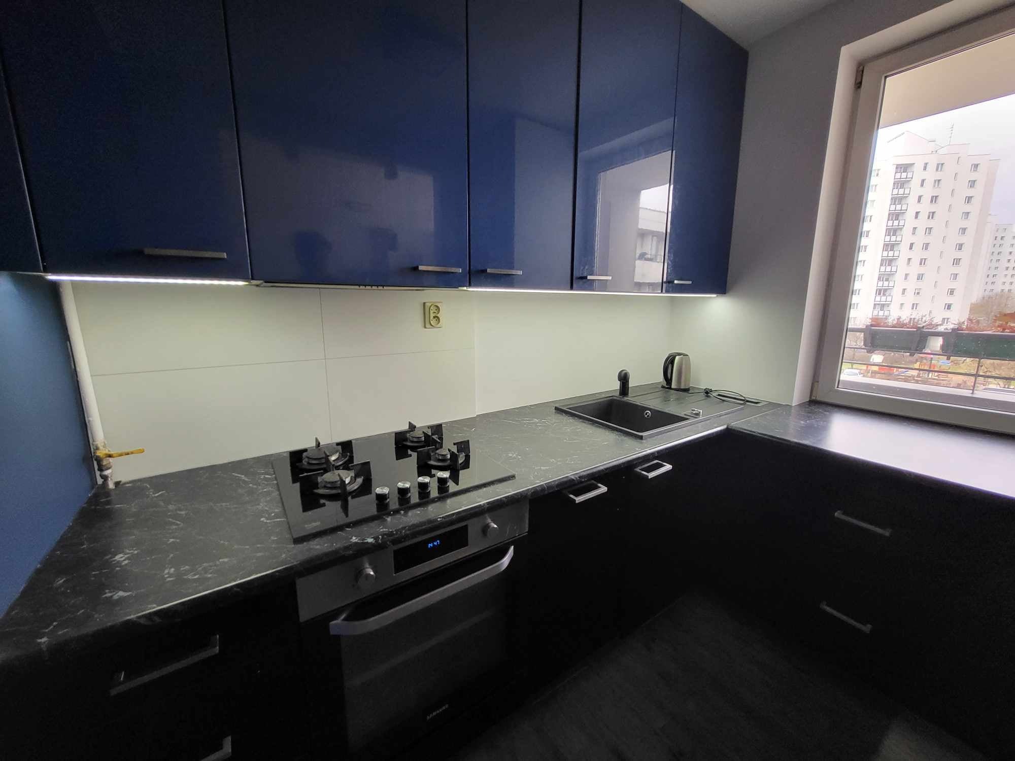 Nowoczesna kuchnia z ciemnymi szafkami i blatem z efektem marmuru, oświetlenie LED pod szafkami, widok na budynek z okna.