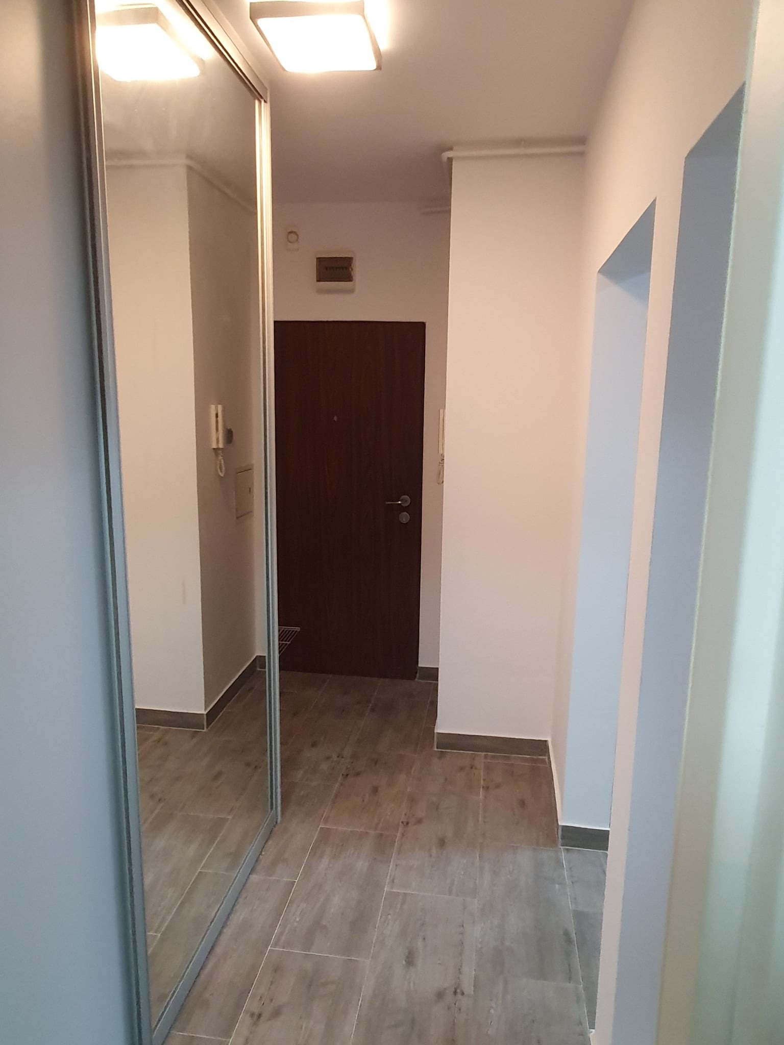 Jasny korytarz po remoncie z przesuwną szafą z lustrem, brązowe drzwi wejściowe i płytki imitujące drewno. Widoczne oświetlenie sufitowe i domofon.