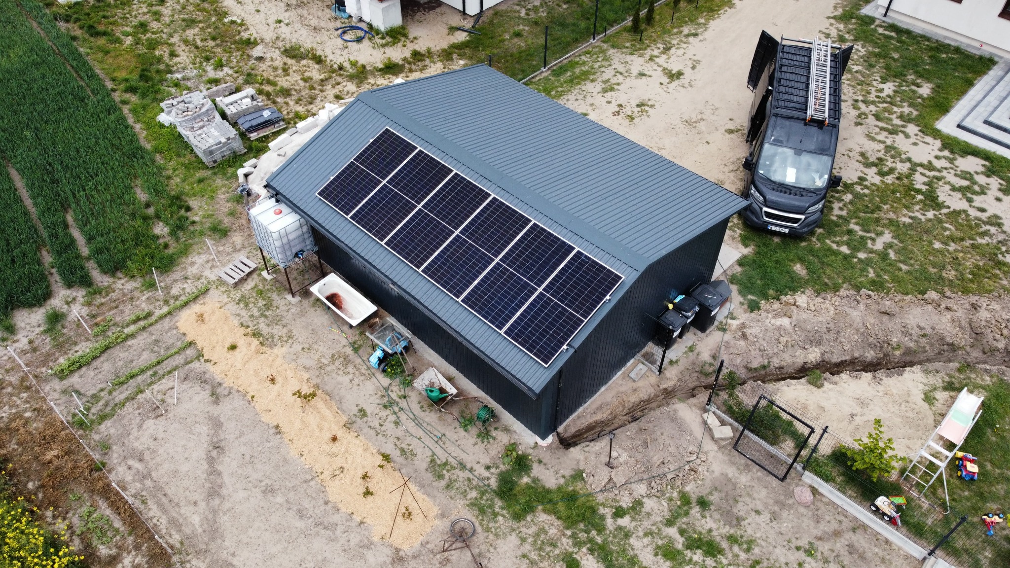 Widok z góry na szary budynek gospodarczy z panelami fotowoltaicznymi na dachu, obok zaparkowany bus z drabiną, w tle pole uprawne i dom.