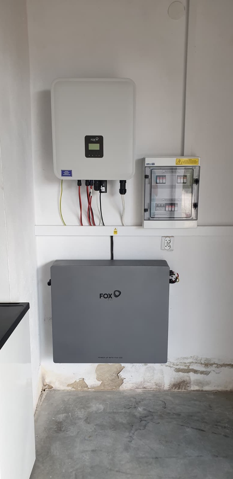 Szary magazyn energii Fox ESS zamontowany na ścianie, obok biały inwerter i skrzynka z bezpiecznikami. Widoczne okablowanie i surowy beton na podłodze.