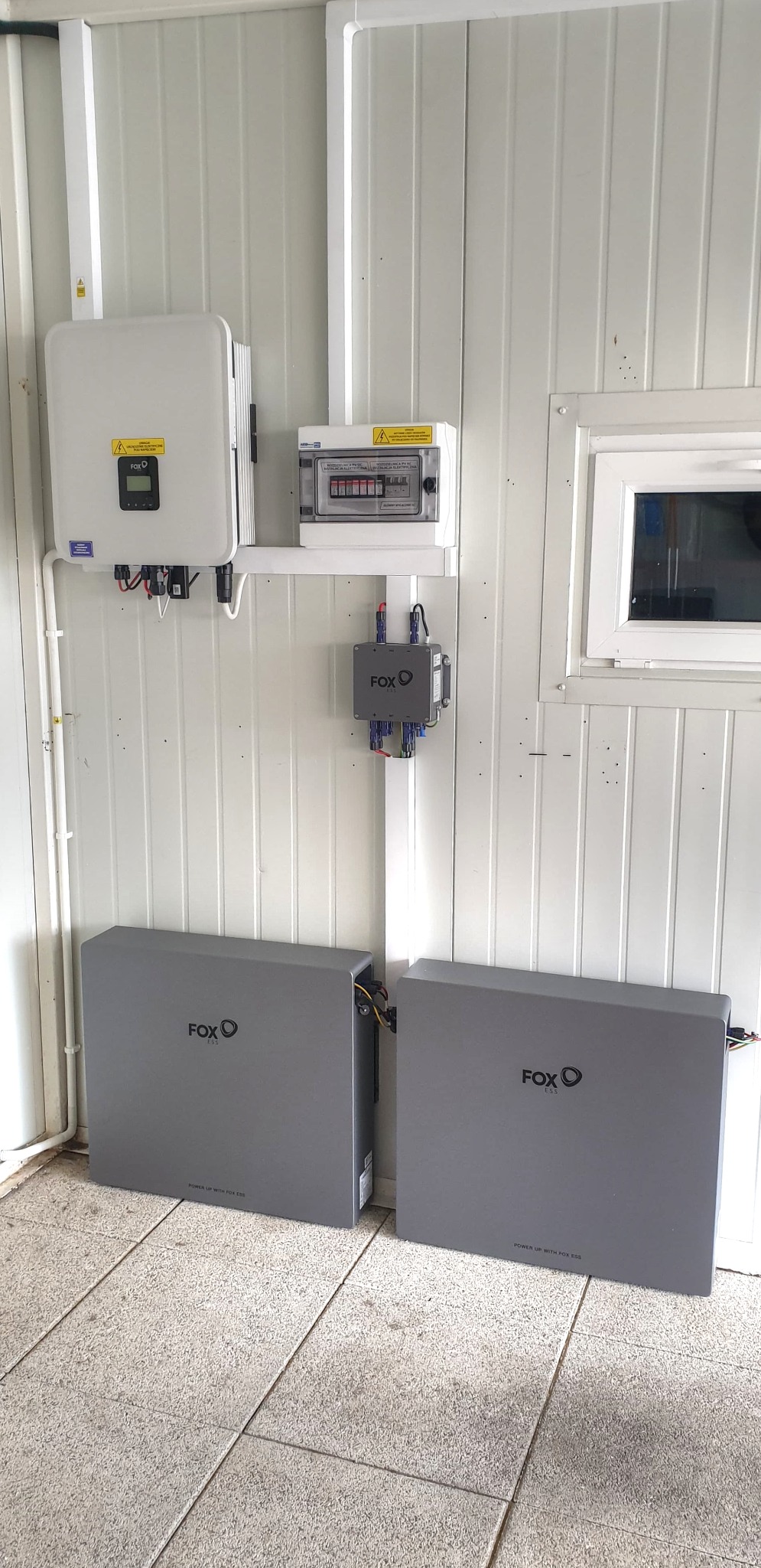 Dwa szare magazyny energii Fox ESS stoją na podłodze w pomieszczeniu z białą, panelową ścianą. Na ścianie zamontowany inwerter, rozdzielnica i moduł komunikacyjny.