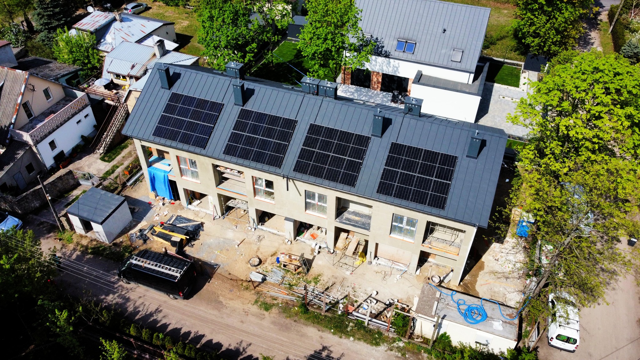 Widok z drona na budynek w trakcie montażu paneli słonecznych na dachu z blachy trapezowej. Na dole widoczne materiały budowlane i samochody dostawcze.