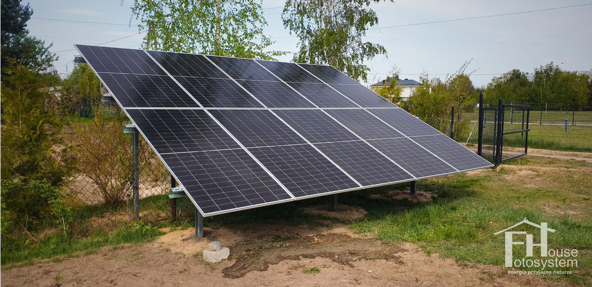 Panel fotowoltaiczny na metalowej konstrukcji, zainstalowany na trawiastym terenie z widocznym ogrodzeniem i zielenią w tle. Ujęcie z niskiej perspektywy.