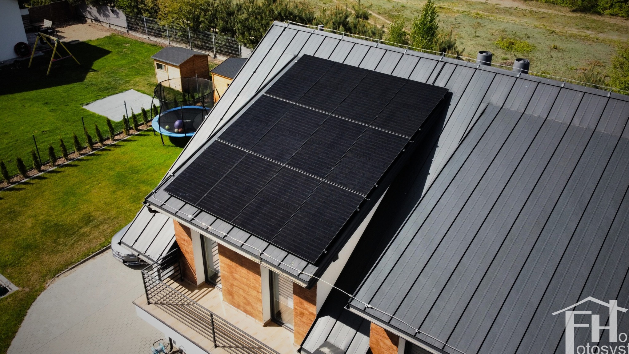 Panele słoneczne na szarym dachu domu jednorodzinnego w Radomiu, widok z góry. W tle zielony trawnik z trampoliną i domkami narzędziowymi.