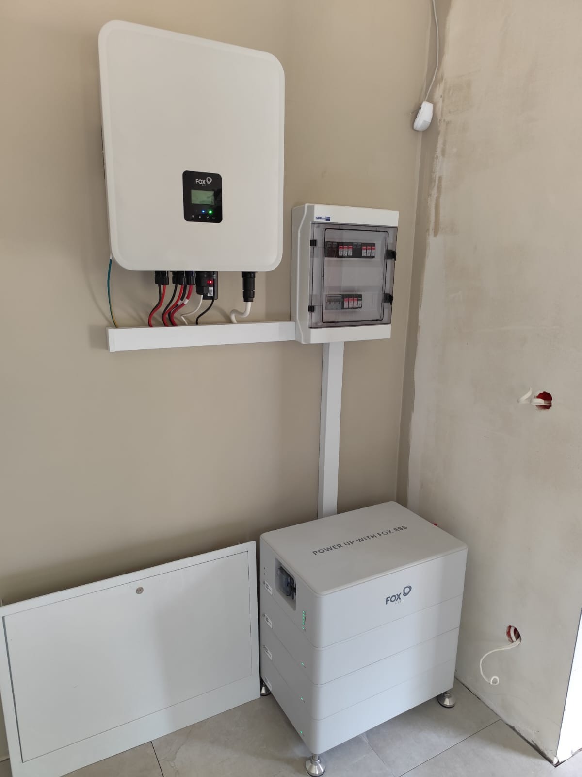 Biały inwerter Fox, skrzynka elektryczna i magazyn energii w jasnym pomieszczeniu. Widoczne kable i elementy instalacji fotowoltaicznej. Ściany w trakcie wykończenia.