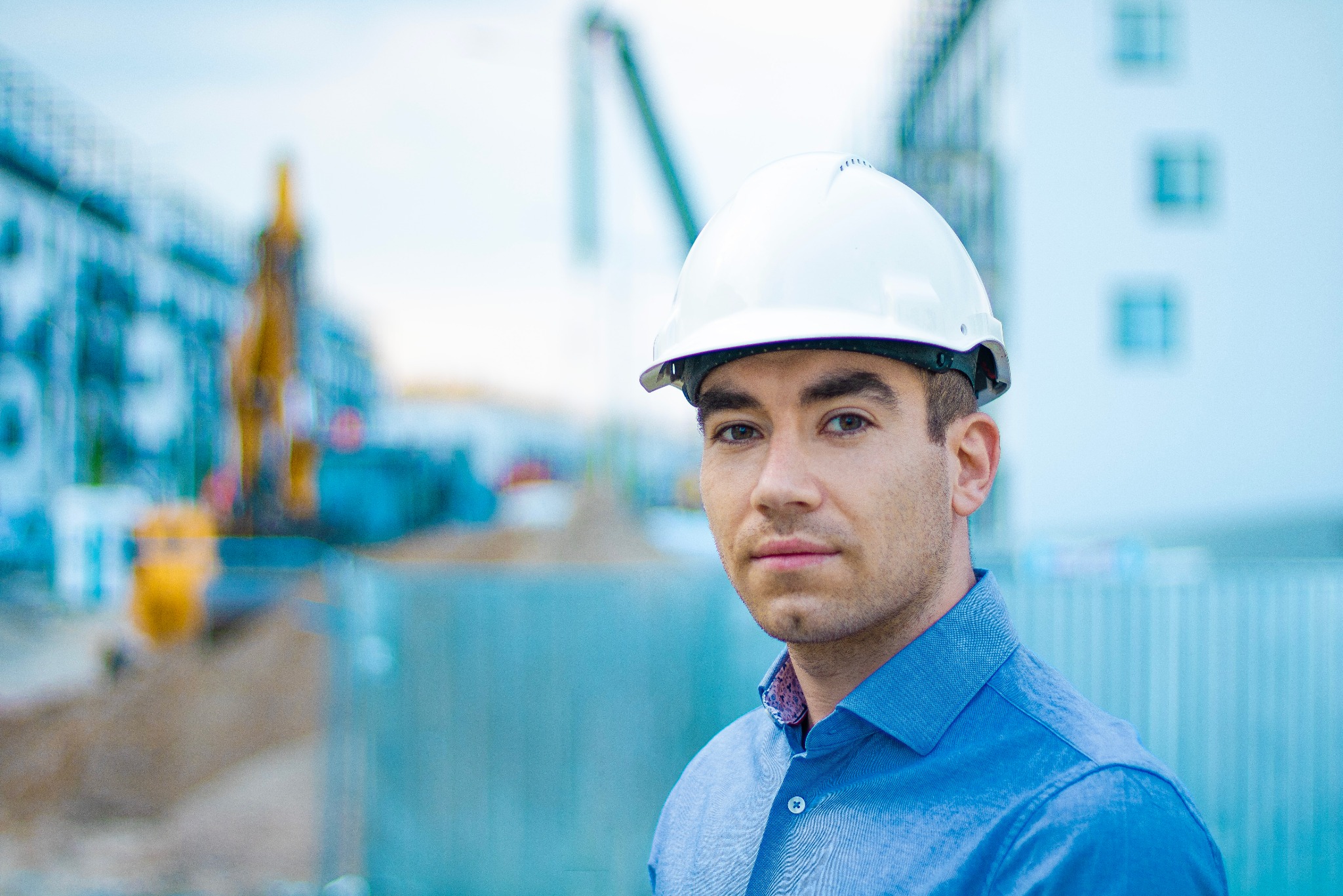 Portret młodego kierownika budowy w białym kasku na tle placu budowy z koparką i powstającym budynkiem. Ujęcie z bliska, skupione na twarzy.