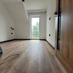 Gwinbud - Jasne poddasze po remoncie: drewniana podłoga, białe ściany z czarnymi akcentami, okno balkonowe z widokiem na zieleń. Widoczny fragment drzwi i sprzęt budowlany.