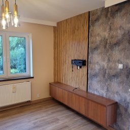 Gwinbud - Nowoczesne wykończenie wnętrza: drewniana zabudowa TV z lamelami i dekoracyjną ścianą, wisząca szafka RTV, parkiet, okno z widokiem na zieleń i designerska lampa.