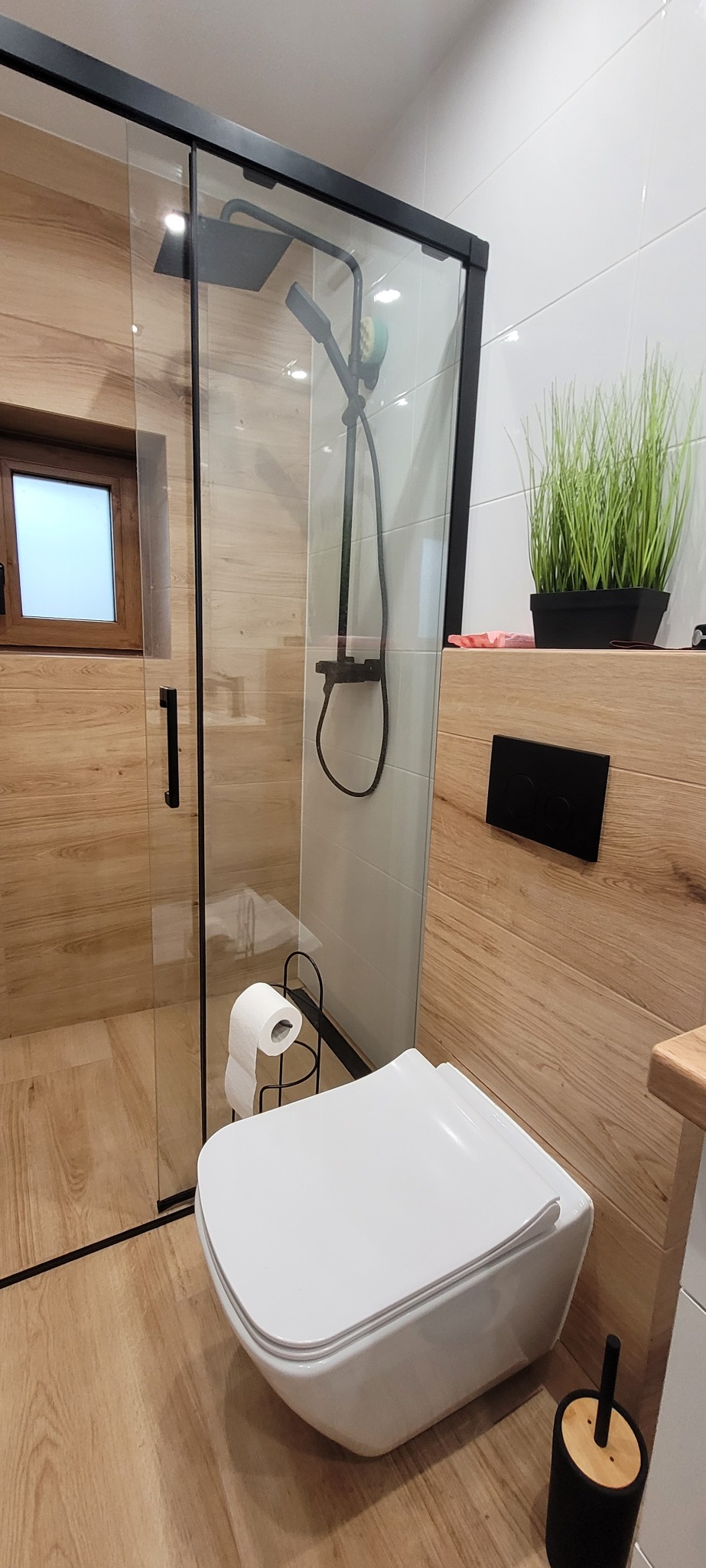 Nowoczesna łazienka z kabiną prysznicową typu walk-in, drewnopodobnymi płytkami i podwieszaną toaletą. Minimalistyczny design z akcentami czerni.