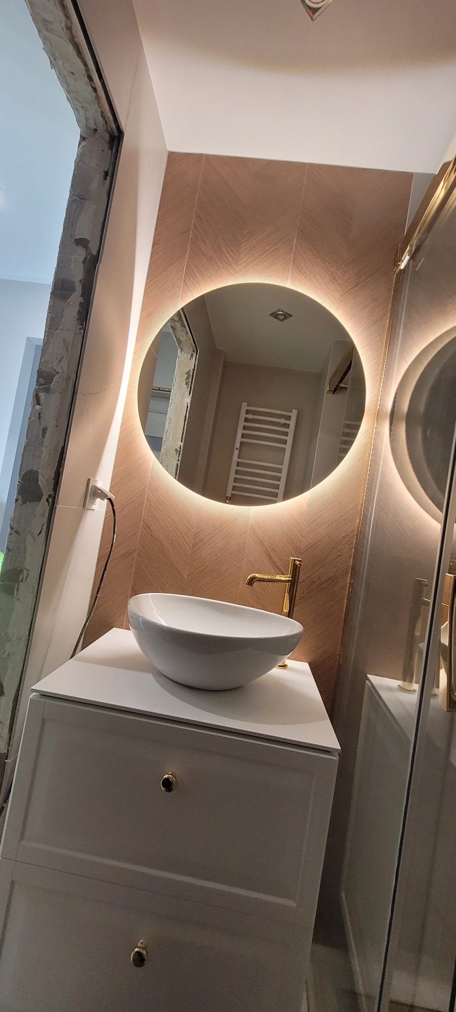 Nowoczesna łazienka w trakcie wykończenia: umywalka nablatowa, złota armatura, podświetlane lustro owalne i dekoracyjna ściana w jodełkę. Widoczne niedokończone obramowanie drzwi.