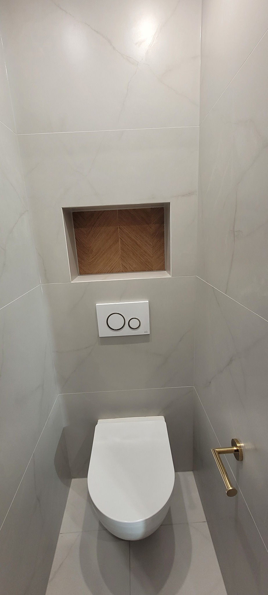 Nowoczesna toaleta w wykończonej łazience z szarymi płytkami imitującymi marmur, drewnianą wnęką i złotym uchwytem na papier toaletowy.