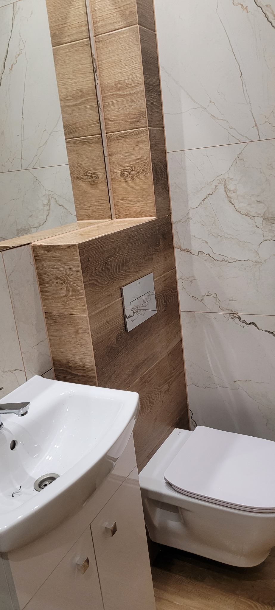 Nowoczesna łazienka po remoncie: biała umywalka z szafką, podwieszana toaleta i marmurowe płytki z drewnianymi akcentami. Minimalistyczny design i funkcjonalność.