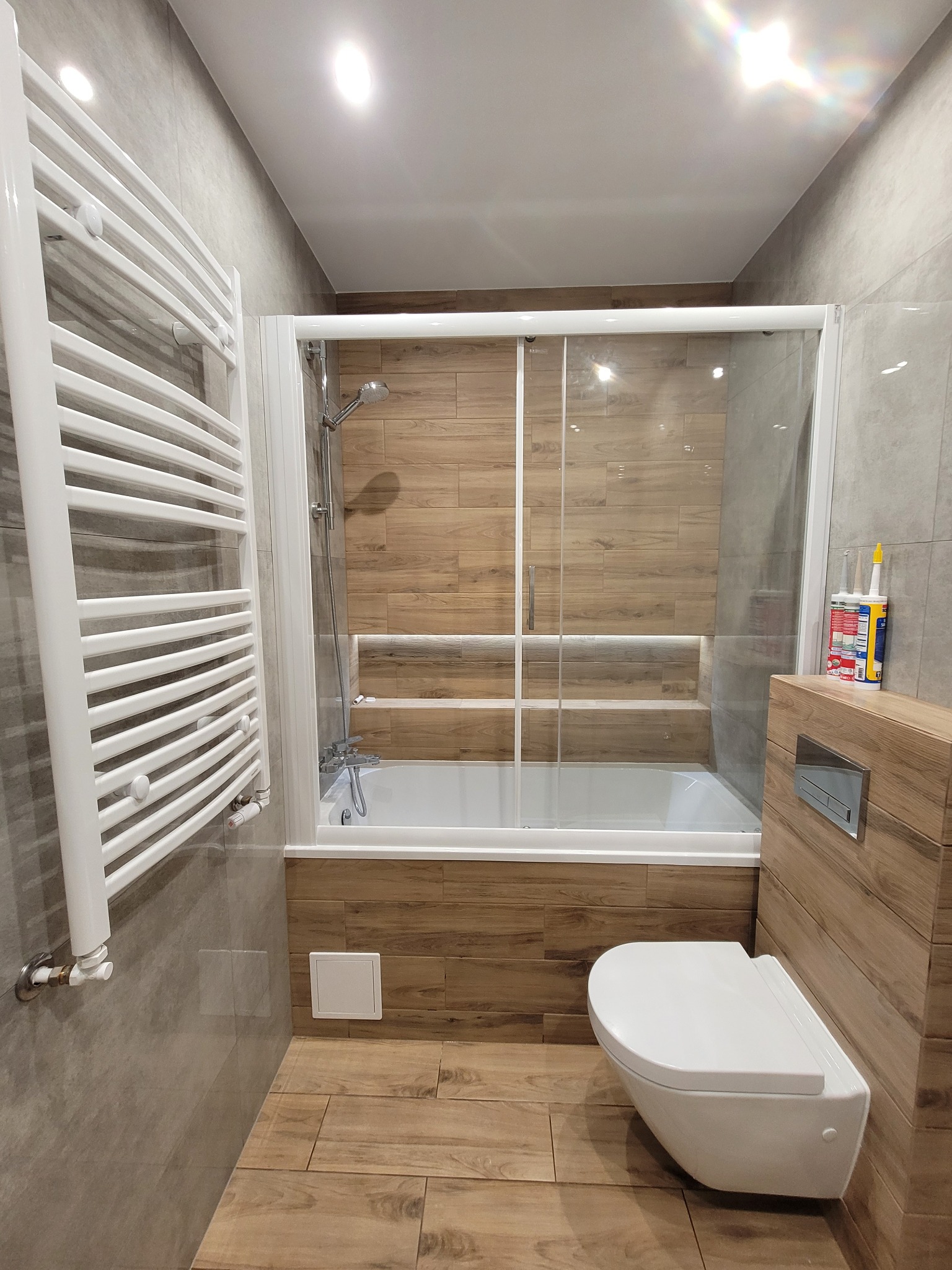 Wykończona łazienka z wanną za przesuwną szybą, wiszącą toaletą i grzejnikiem drabinkowym. Płytki imitujące drewno na podłodze i ścianie w strefie kąpieli.