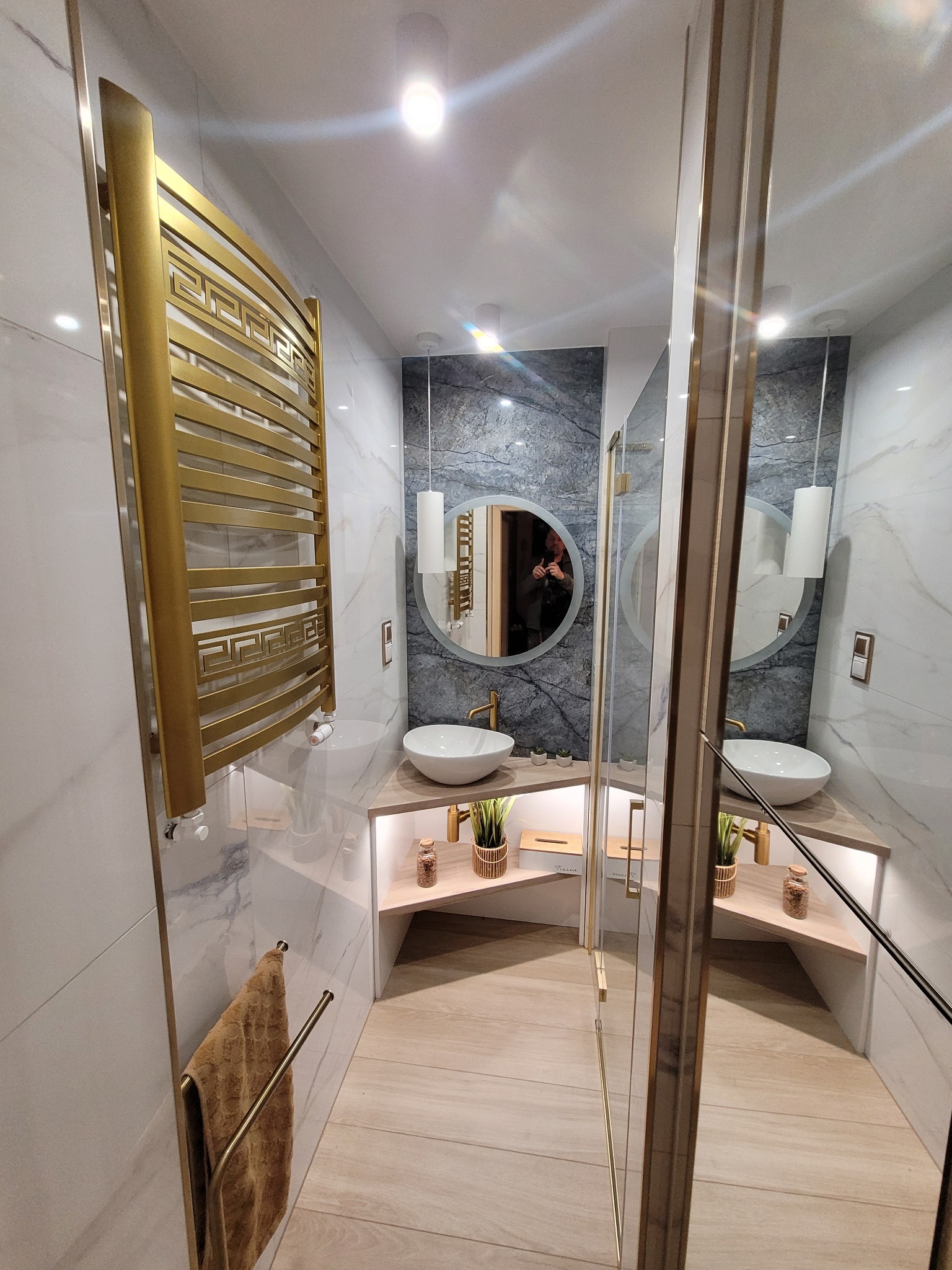 Elegancka łazienka po remoncie: marmurowe ściany, złoty grzejnik drabinkowy, okrągłe lustro i kabina prysznicowa ze złotymi detalami. Nowoczesny design.
