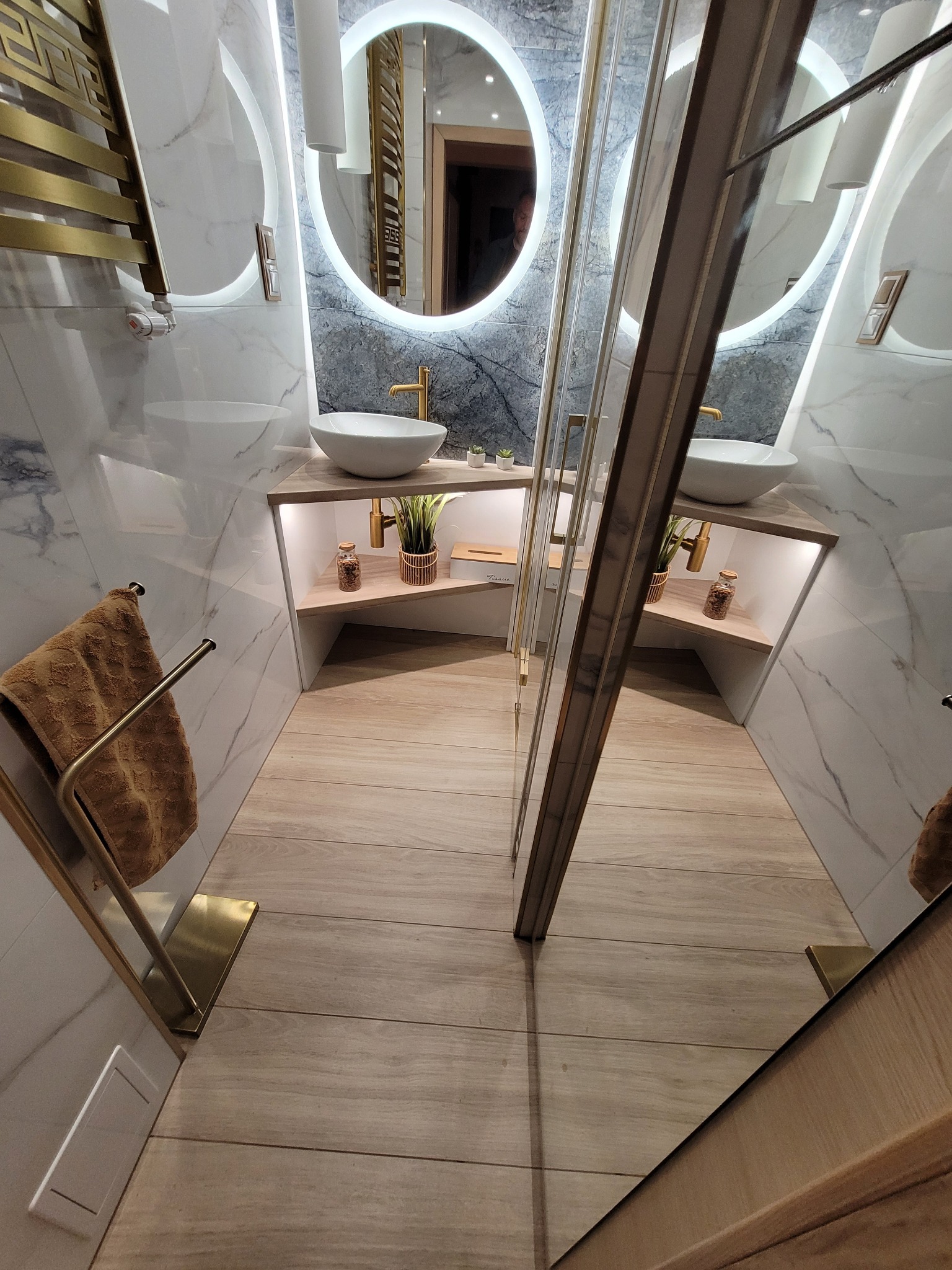 Nowoczesna łazienka z marmurowymi ścianami, okrągłymi lustrami z podświetleniem LED, drewnianą podłogą i złotymi akcentami, w tym armaturą i grzejnikiem.