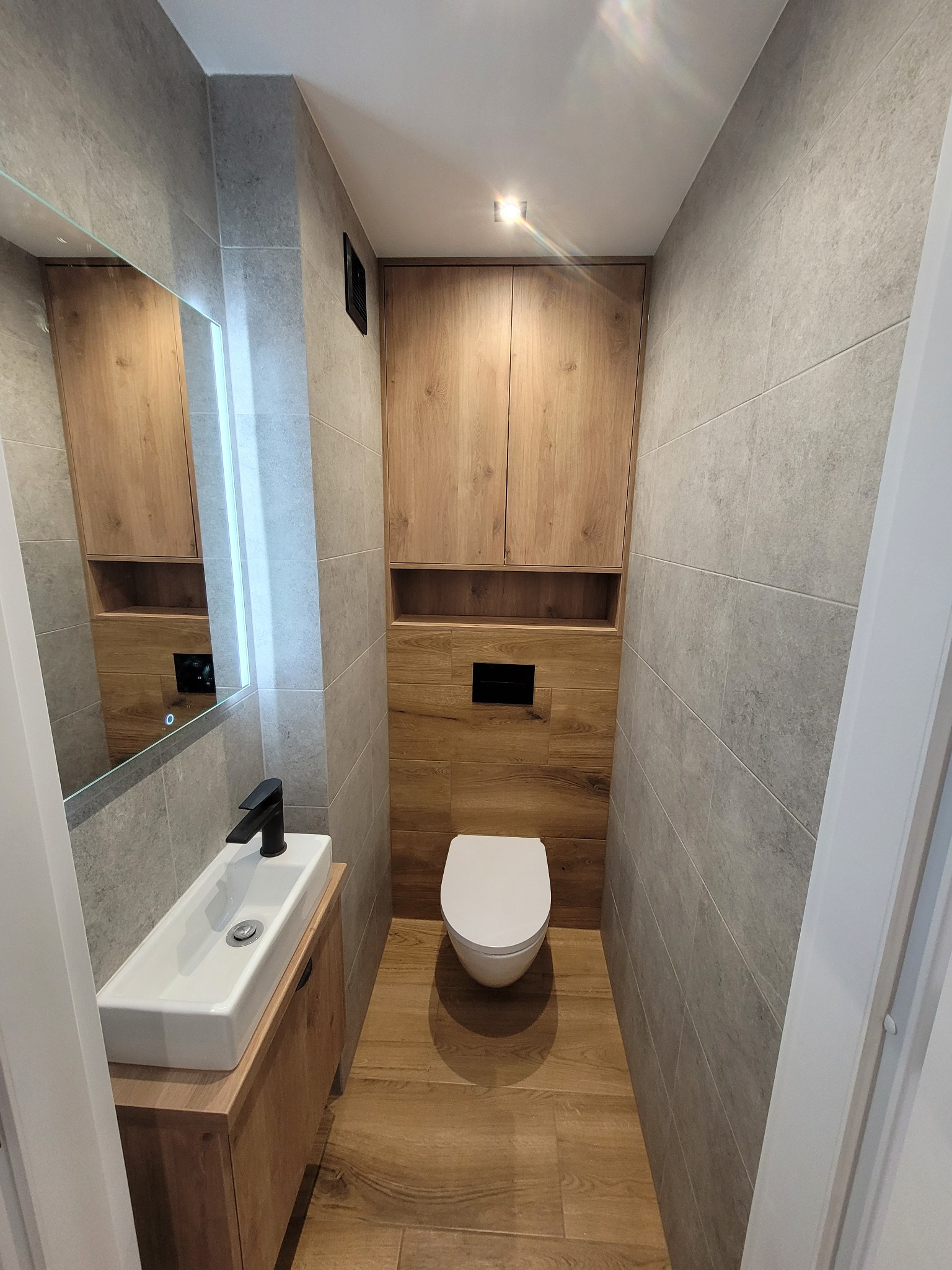 Wąska toaleta w szaro-drewnianej tonacji: szare płytki ścienne, drewnopodobna podłoga, wisząca szafka nad WC i umywalka z szafką. Nowoczesne wykończenie.