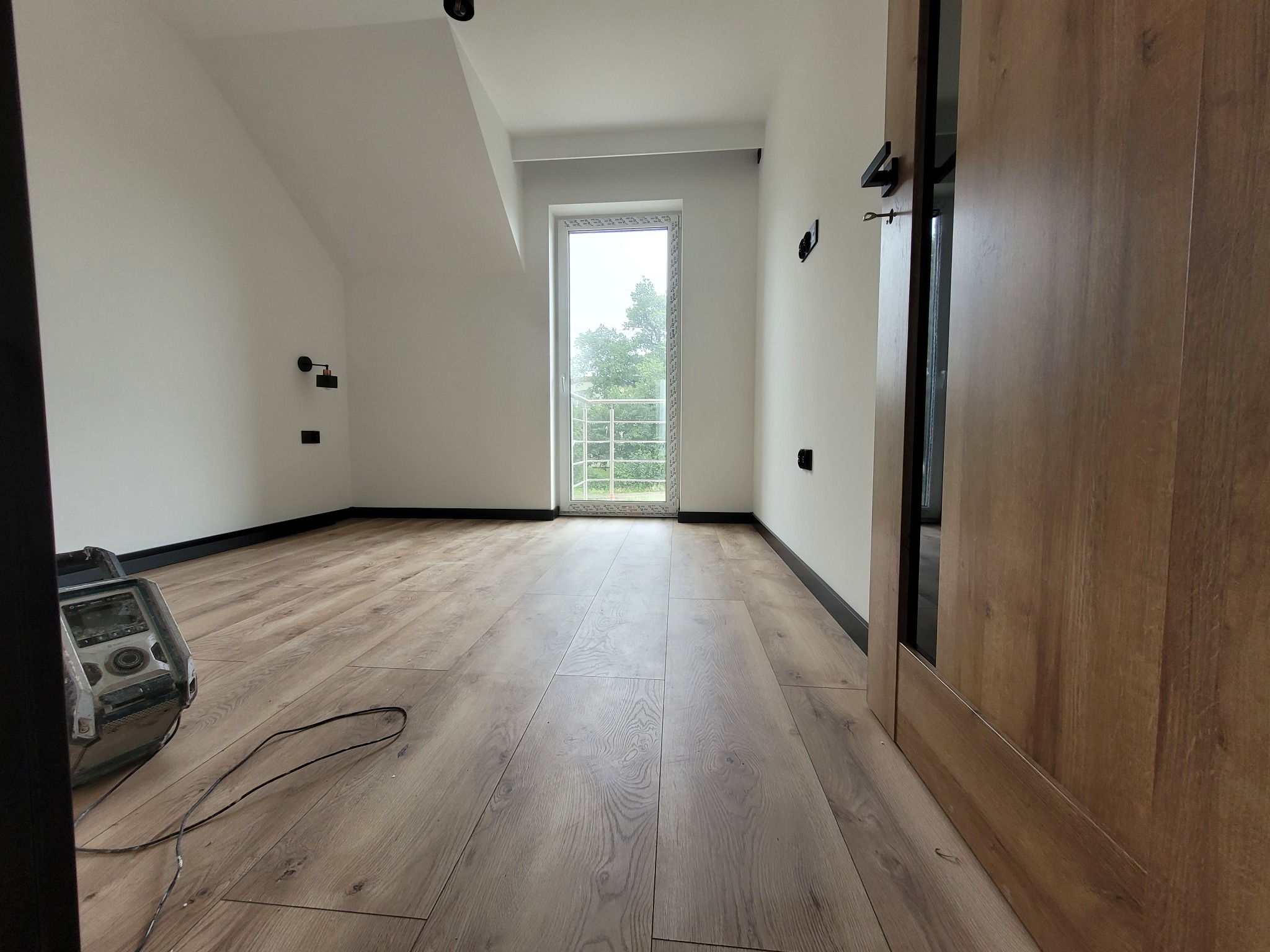 Jasne poddasze po remoncie: drewniana podłoga, białe ściany z czarnymi akcentami, okno balkonowe z widokiem na zieleń. Widoczny fragment drzwi i sprzęt budowlany.