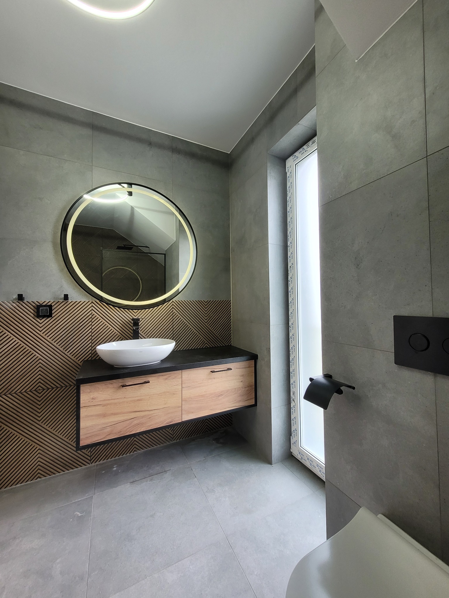 Nowoczesna łazienka z szarą płytką, okrągłym lustrem z podświetleniem LED, drewnianą szafką pod umywalką i dekoracyjną ścianą z geometrycznym wzorem.