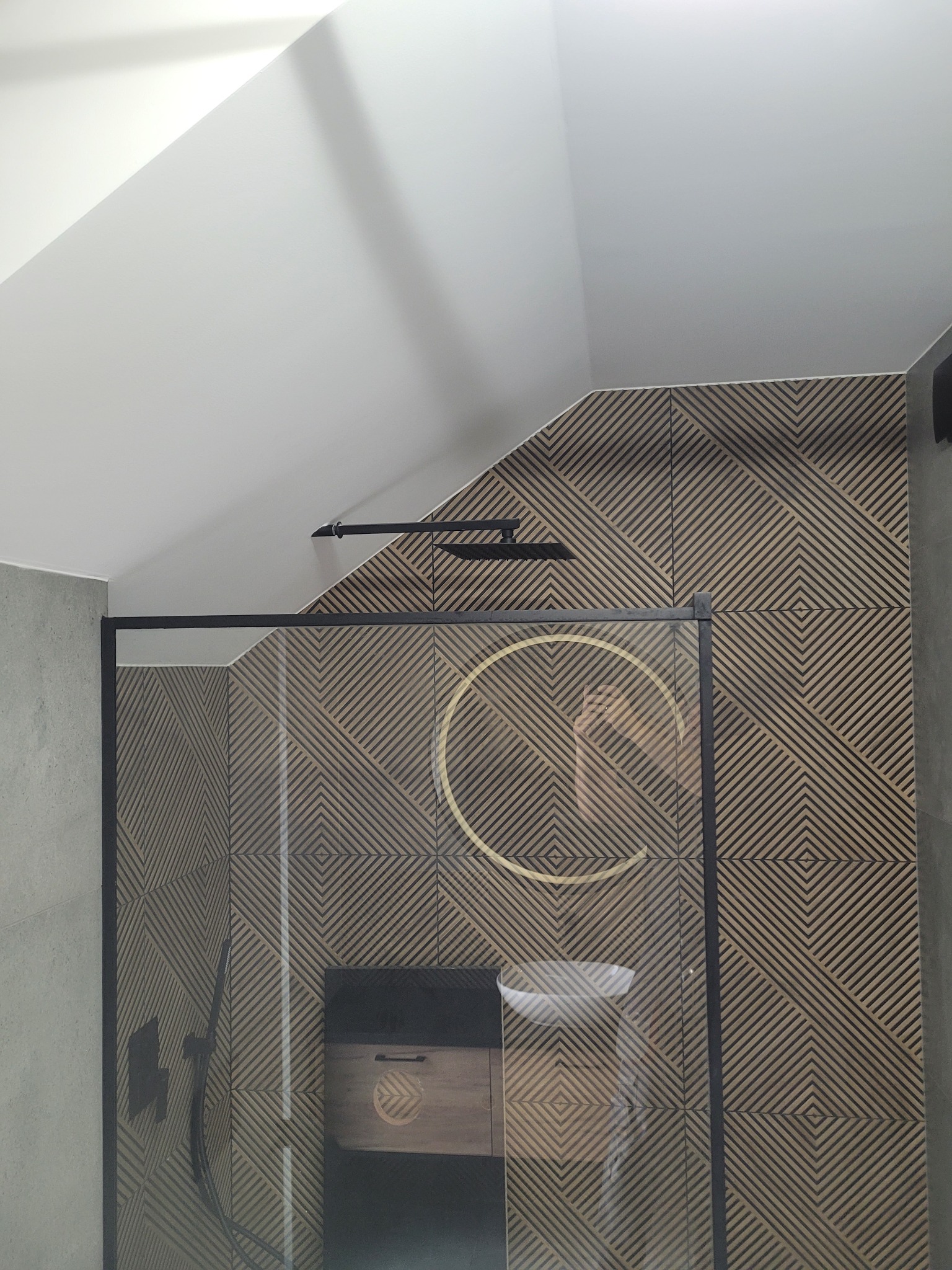 Nowoczesna łazienka na poddaszu z kabiną prysznicową typu walk-in, wykończona płytkami z geometrycznym wzorem i czarną armaturą. Umywalka z szafką wkomponowana w aranżację.