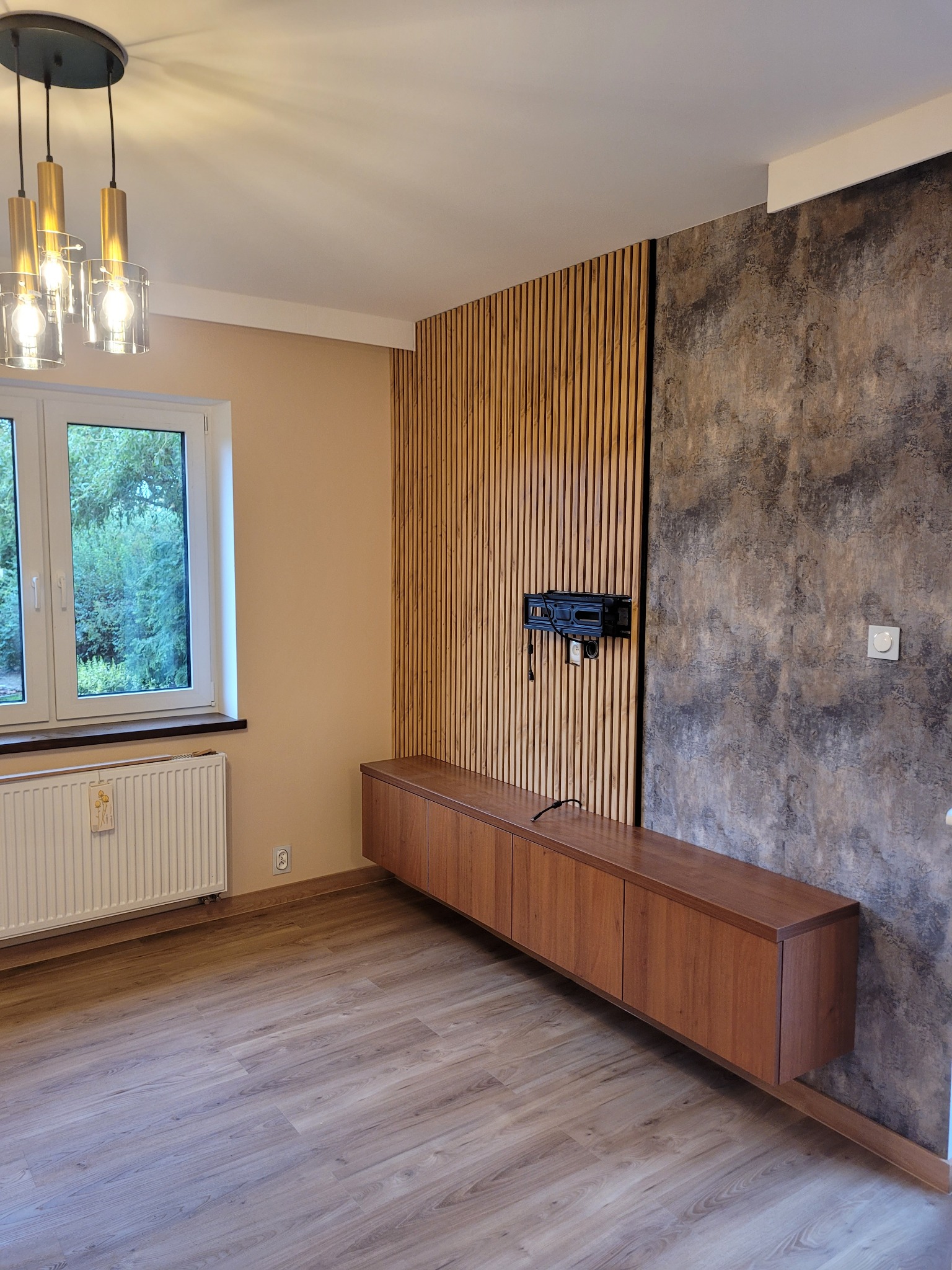Nowoczesne wykończenie wnętrza: drewniana zabudowa TV z lamelami i dekoracyjną ścianą, wisząca szafka RTV, parkiet, okno z widokiem na zieleń i designerska lampa.