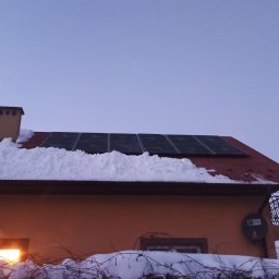 VOLTRI SPÓŁKA Z OGRANICZONĄ ODPOWIEDZIALNOŚCIĄ - Panele fotowoltaiczne na dachu domu pokrytym śniegiem, widoczne oświetlone okno i balkon z ozdobną balustradą. Zimowy krajobraz o zmierzchu.