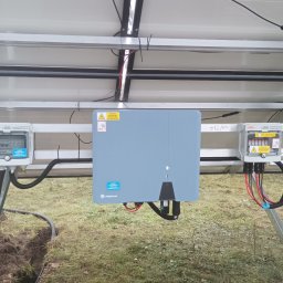 VOLTRI SPÓŁKA Z OGRANICZONĄ ODPOWIEDZIALNOŚCIĄ - Montaż paneli słonecznych z widocznym inwerterem Solplanet i skrzynkami KENO. Instalacja na trawiastym terenie, widok od spodu konstrukcji.