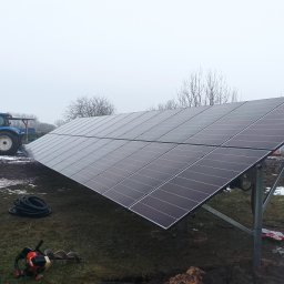 VOLTRI SPÓŁKA Z OGRANICZONĄ ODPOWIEDZIALNOŚCIĄ - Panel fotowoltaiczny na polu uprawnym, z traktorem w tle. Widoczne narzędzia i akcesoria montażowe. Pochmurny dzień, instalacja w trakcie prac.