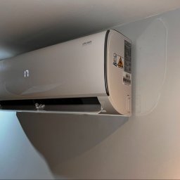 VOLTRI SPÓŁKA Z OGRANICZONĄ ODPOWIEDZIALNOŚCIĄ - Nowoczesny, biały klimatyzator ścienny Ukura Series z cyfrowym wyświetlaczem '17' i przezroczystym deflektorem, zamontowany na jasnej ścianie.