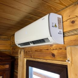 VOLTRI SPÓŁKA Z OGRANICZONĄ ODPOWIEDZIALNOŚCIĄ - Biały klimatyzator ścienny UKURA z cyfrowym wyświetlaczem '19', zamontowany na rustykalnej drewnianej ścianie z widocznymi słojami. W tle fragment okna i rzeźbionego drewnianego mebla.
