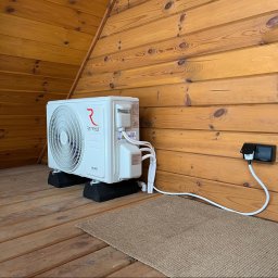 VOLTRI SPÓŁKA Z OGRANICZONĄ ODPOWIEDZIALNOŚCIĄ - Biała jednostka zewnętrzna klimatyzacji Rotenso Ukura na czarnych podkładkach, zainstalowana na drewnianej podłodze przy skośnej ścianie. Kabel podłączony do gniazdka.