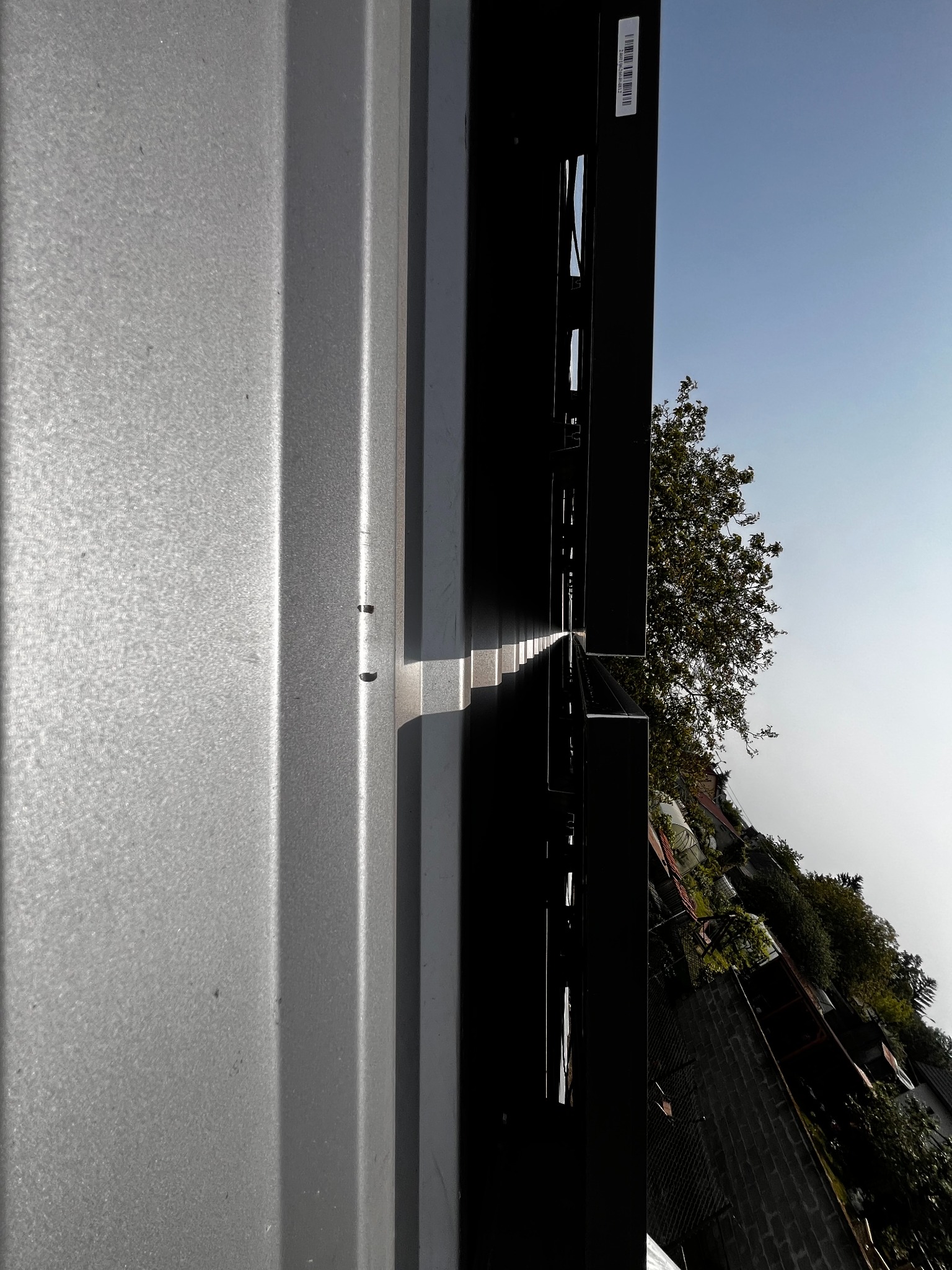 Szara, metalowa konstrukcja z widocznymi elementami montażowymi i fragmentem nieba w tle. Widoczne detale mocowania i faktura blachy. Ujęcie z dołu, podkreślające pionowość.