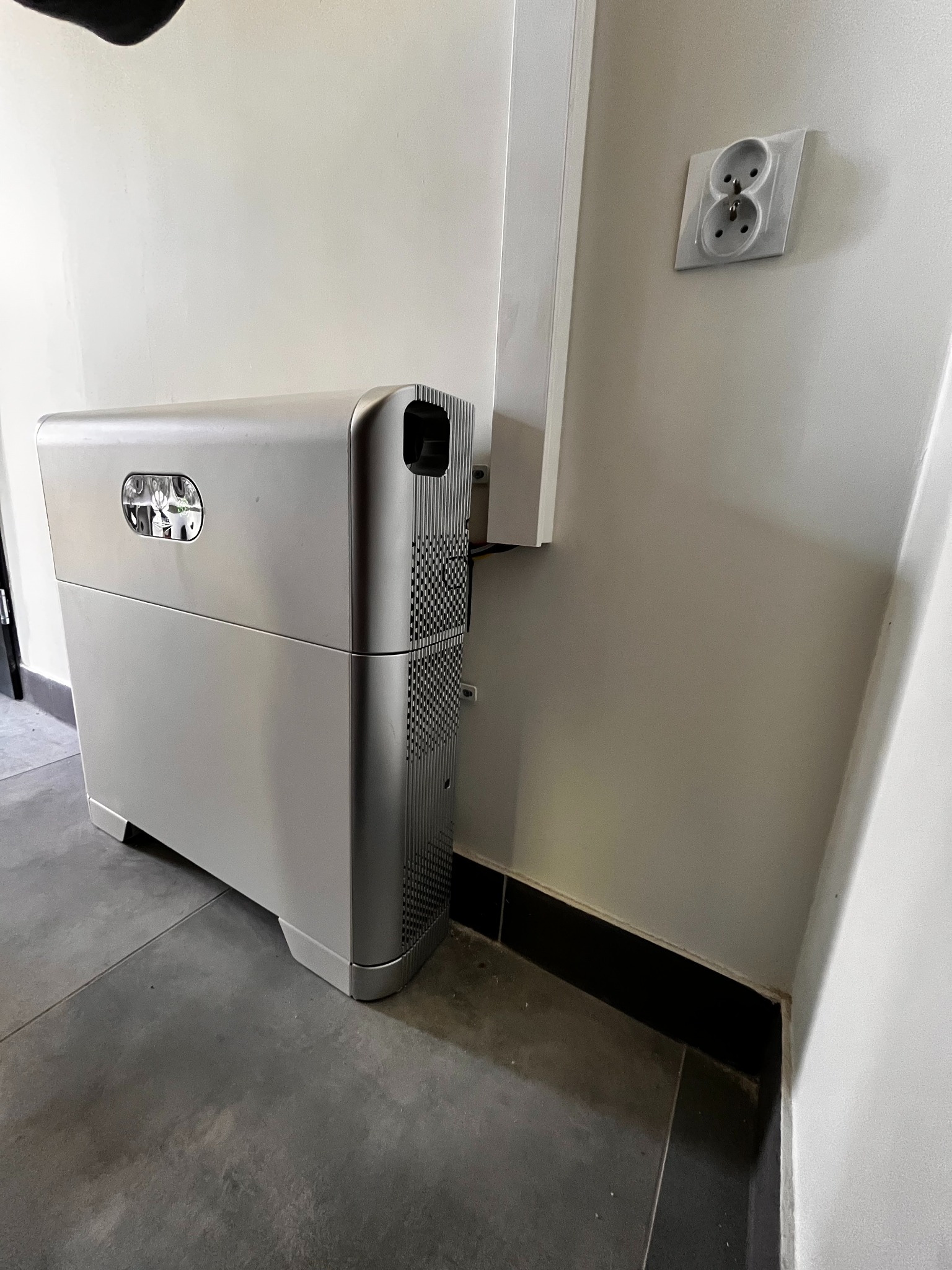 Szary magazyn energii w pomieszczeniu, widoczny fragment instalacji elektrycznej i gniazdko. Minimalistyczny design urządzenia, nowoczesne wykończenie wnętrza.
