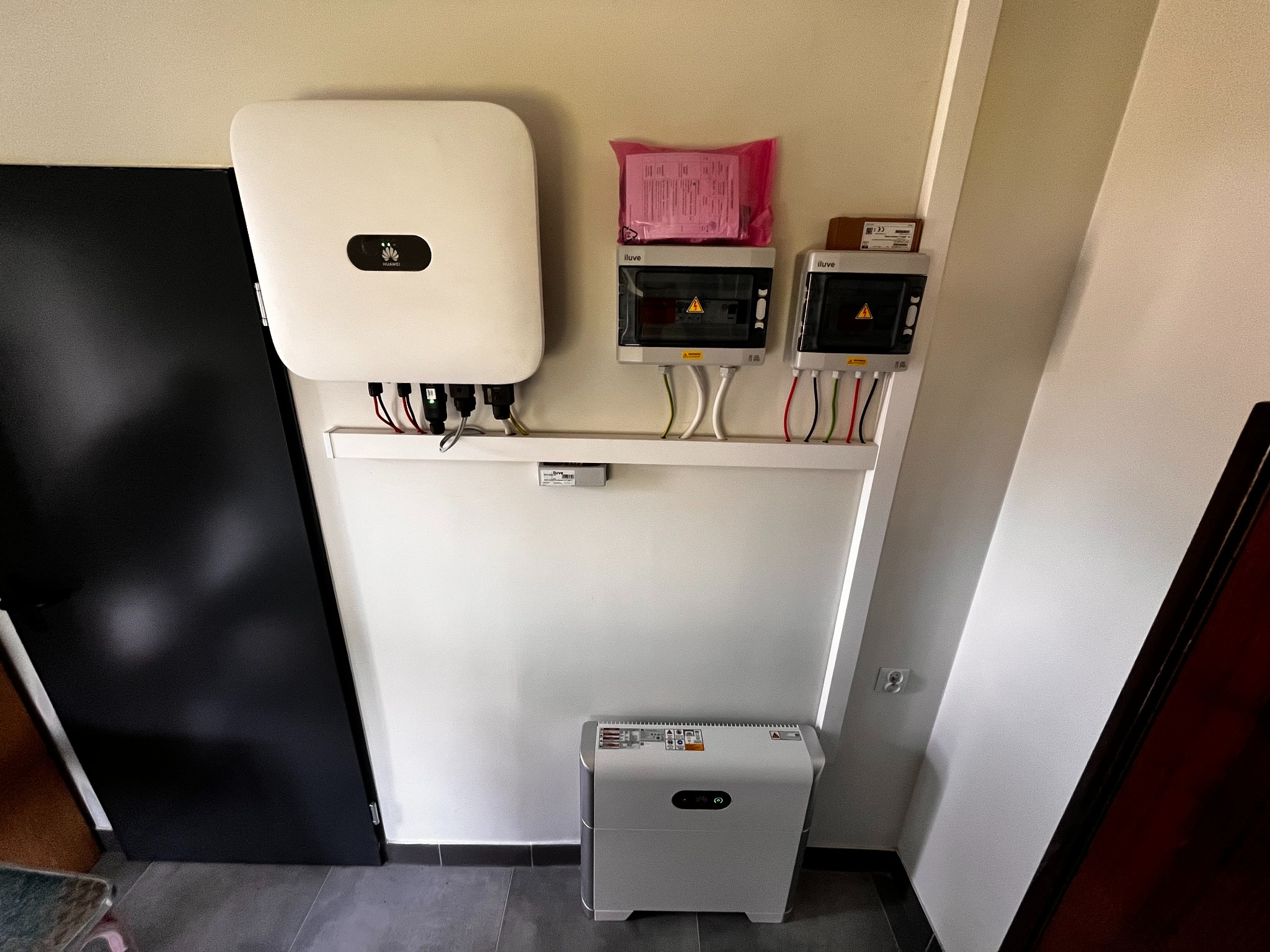 Biały inwerter Huawei, dwa rozdzielacze elektryczne i magazyn energii na ścianie wewnątrz pomieszczenia. Widoczne przewody i elementy instalacji.