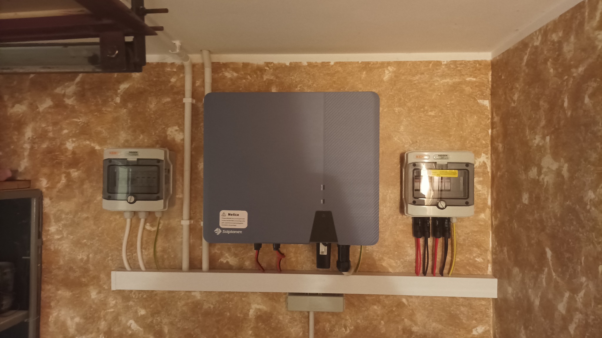Szary falownik Solplanet zamontowany na ścianie o teksturze, z dwoma skrzynkami przyłączeniowymi po bokach. Widoczne przewody i rurki instalacyjne.
