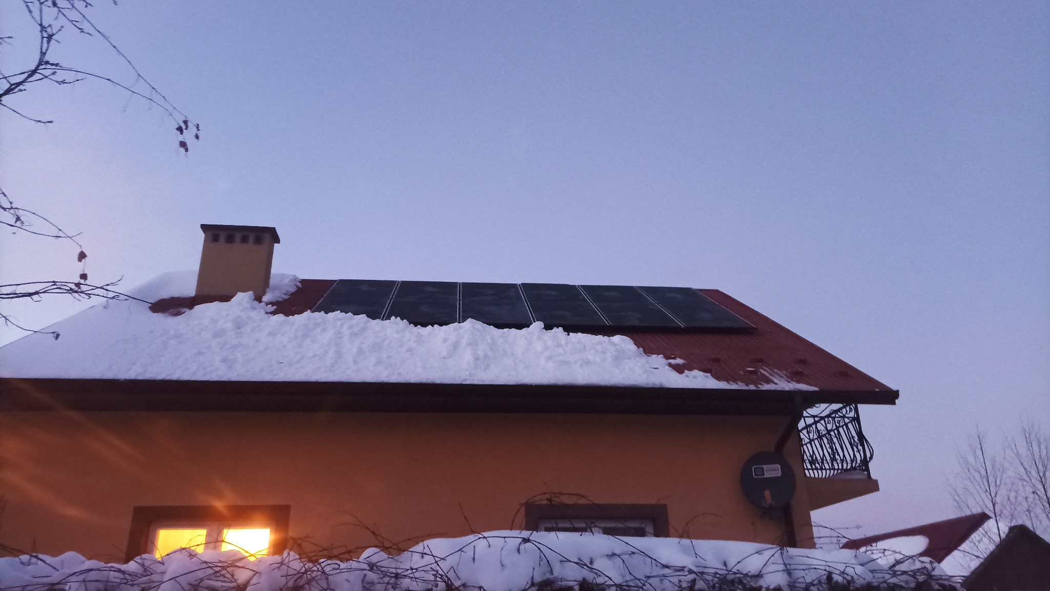 Panele fotowoltaiczne na dachu domu pokrytym śniegiem, widoczne oświetlone okno i balkon z ozdobną balustradą. Zimowy krajobraz o zmierzchu.