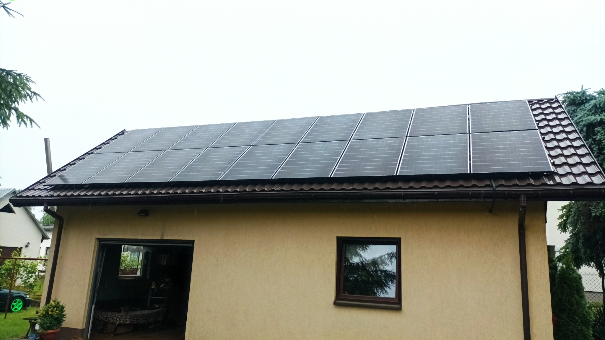 Panele słoneczne na dachu budynku z beżową elewacją, widoczne okno i brama garażowa. Ujęcie z dołu, pochmurna pogoda.