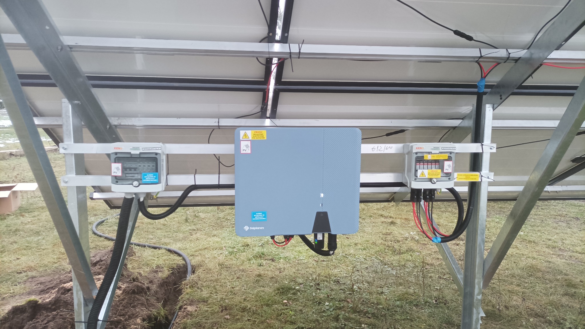 Montaż paneli słonecznych z widocznym inwerterem Solplanet i skrzynkami KENO. Instalacja na trawiastym terenie, widok od spodu konstrukcji.