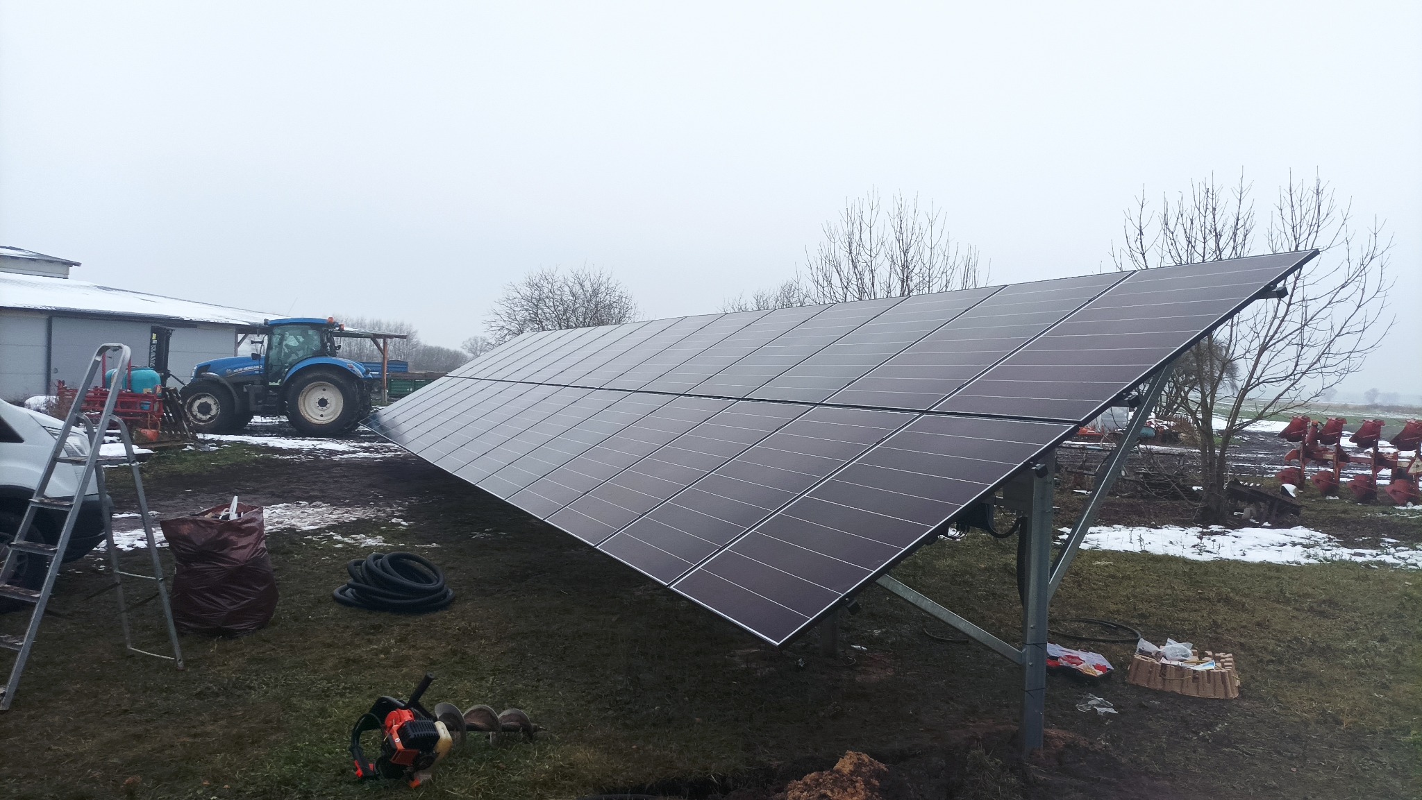 Panel fotowoltaiczny na polu uprawnym, z traktorem w tle. Widoczne narzędzia i akcesoria montażowe. Pochmurny dzień, instalacja w trakcie prac.