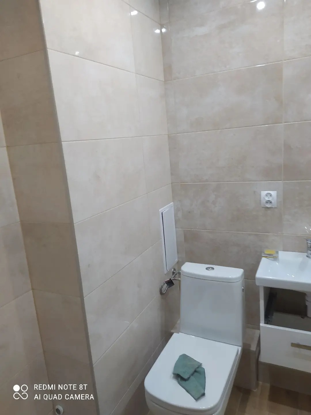 Jasna łazienka po remoncie: beżowe płytki na ścianach, biała toaleta z zielonym ręcznikiem, umywalka z szafką. Widoczny fragment ściany z dostępem do instalacji.