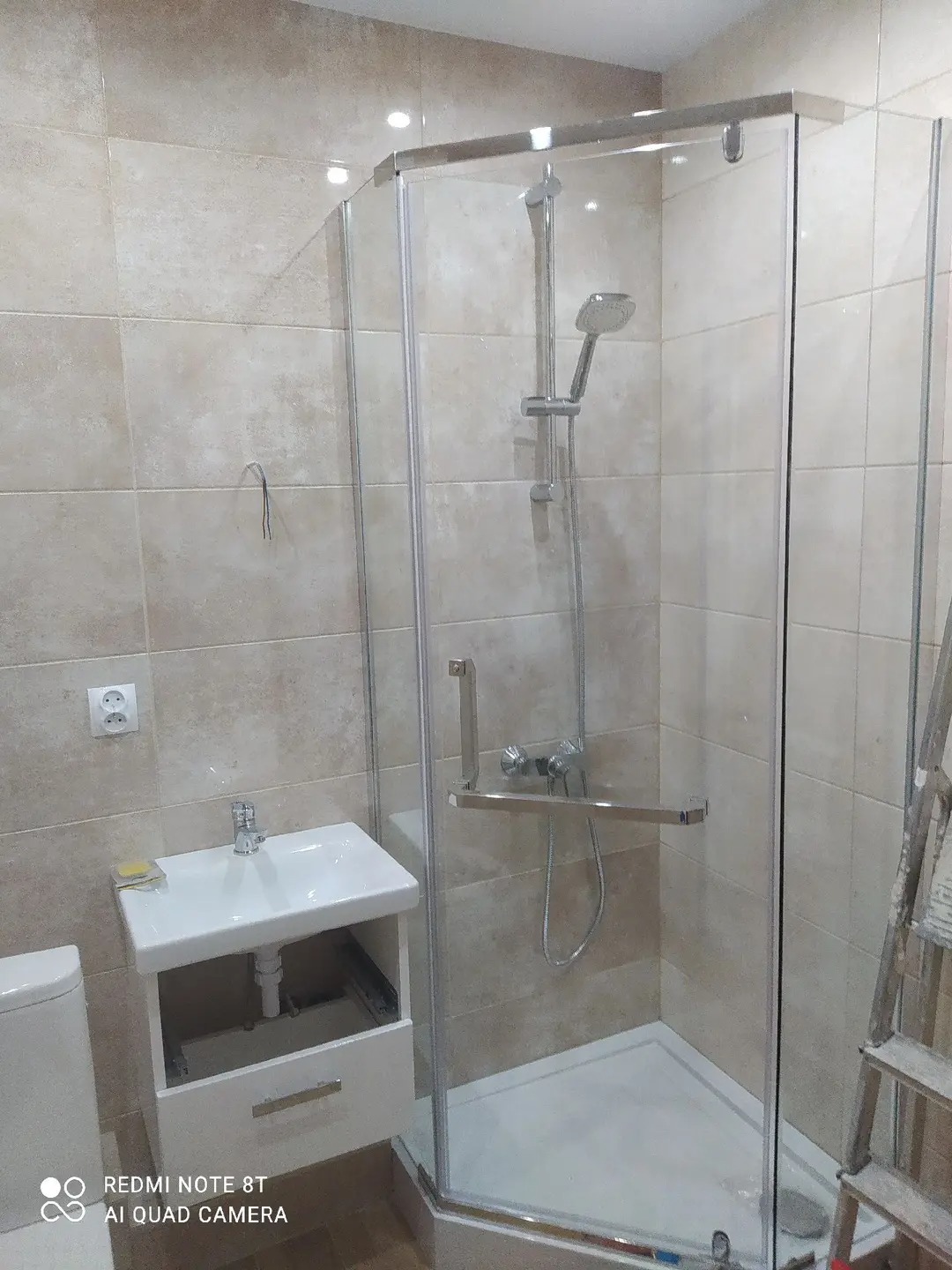 Wykończona łazienka z kabiną prysznicową narożną, umywalką z szafką i beżowymi płytkami. Widoczna drabina malarska. Nowoczesny design.