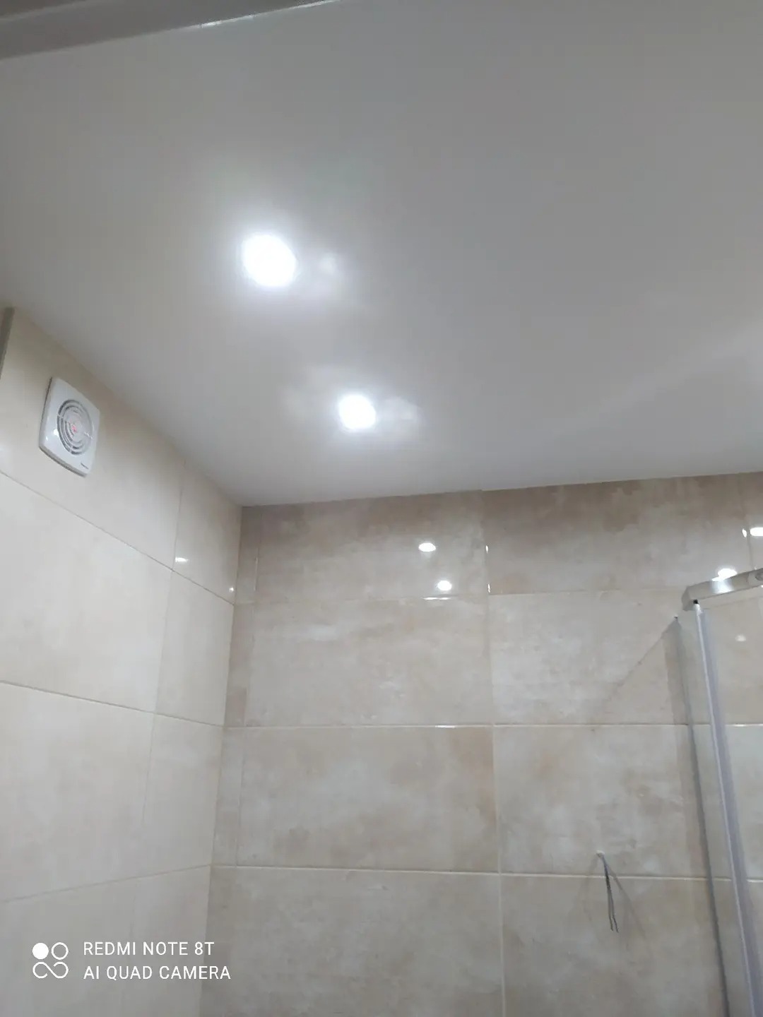 Wykończona łazienka z beżowymi płytkami, widoczne oświetlenie sufitowe i wentylator. Fragment kabiny prysznicowej po prawej stronie. Ujęcie z dołu, podkreślające detale wykończenia.