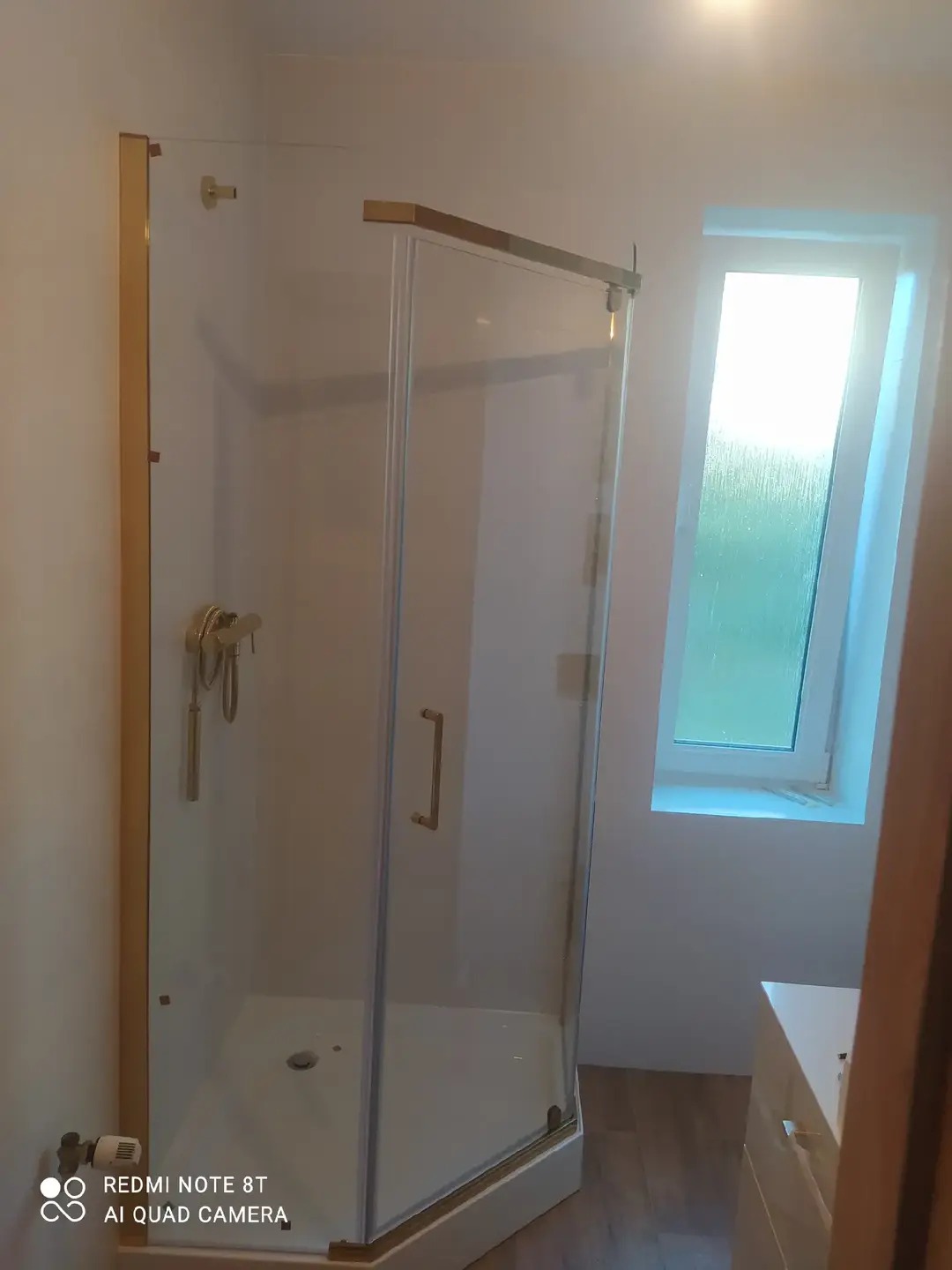 Nowoczesna kabina prysznicowa z elementami w kolorze złotym, widoczna w jasnej łazience z oknem. Ukończony montaż, czyste wykończenie.