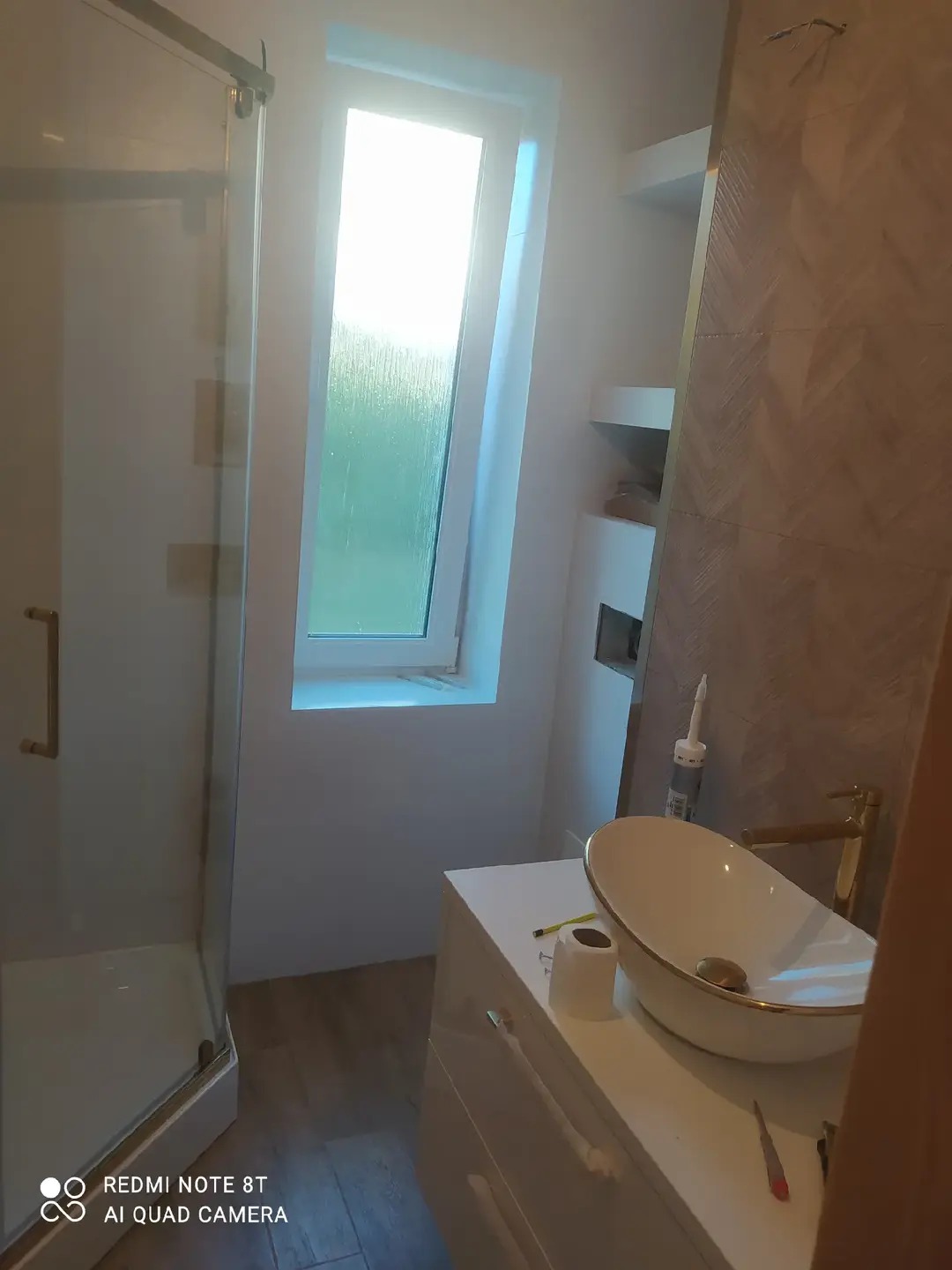 Nowoczesna łazienka po remoncie: kabina prysznicowa ze złotymi detalami, umywalka nablatowa na białej szafce, okno w skosie i dekoracyjna ściana z płytek.