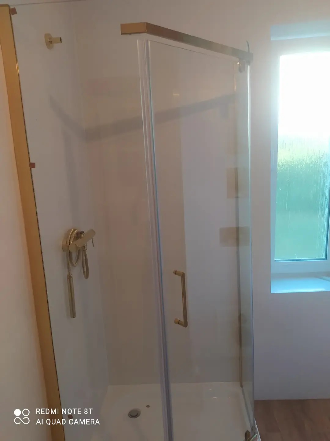 Nowoczesna kabina prysznicowa z złotymi detalami, szklane drzwi, widoczny odpływ i bateria. W tle okno z widokiem na zieleń. Minimalistyczny design.