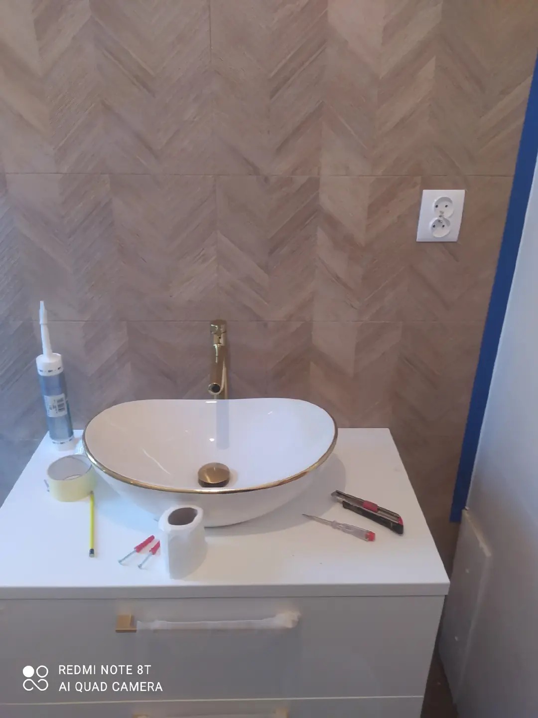 Biała umywalka nablatowa z złotymi akcentami na szafce w łazience z płytkami ułożonymi w jodełkę. Widoczne narzędzia i materiały montażowe.